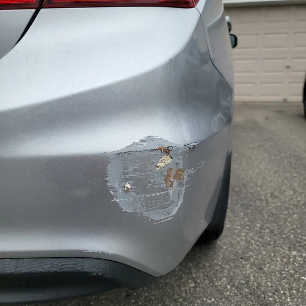 Curb-rash scuff on the lower rear corner of a silver car bumper, paint scraped off exposing primer, photographed in a driveway