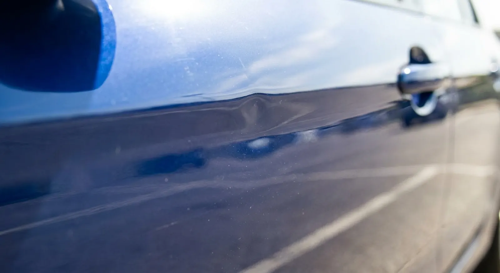 Close-up of a parking-lot door ding and shallow crease dent on a glossy blue car door panel