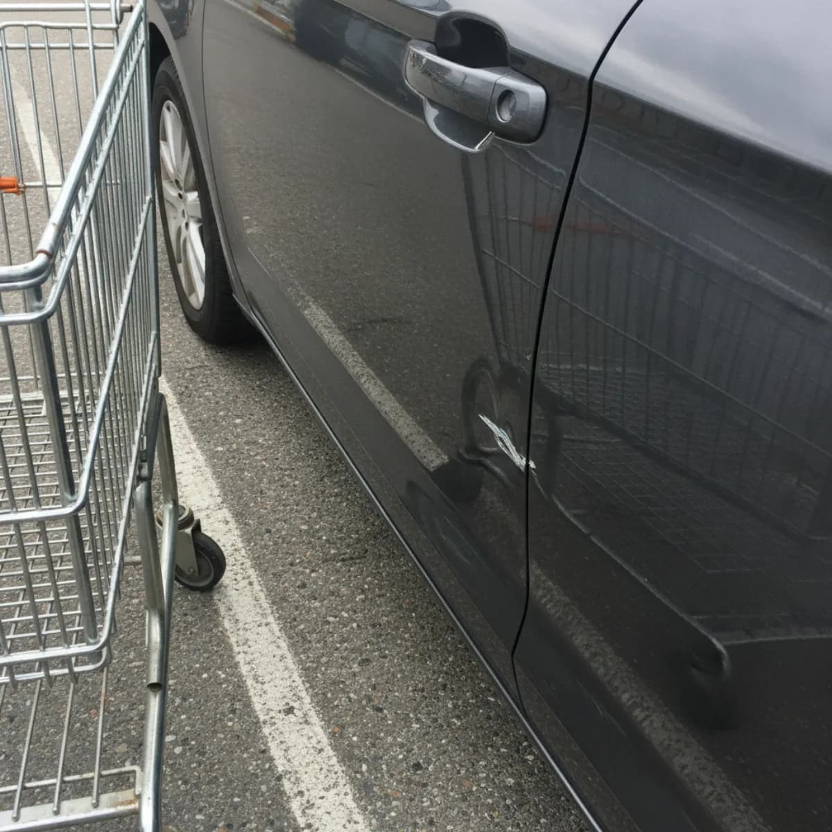 A chrome wire shopping cart parked close alongside a dark grey sedan in a supermarket lot, with a small light-coloured dent and paint scuff low on the car's rear door and the sky's reflection breaking across the damaged spot