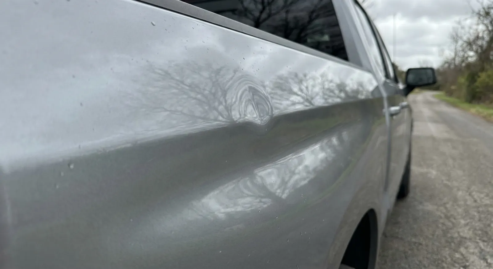 Close-up of a shallow dent on the silver tailgate of a pickup truck, trees reflected across the panel